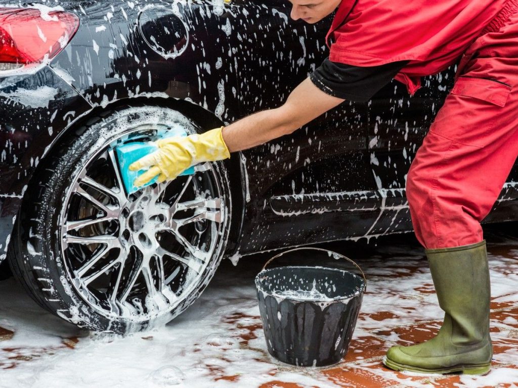 Car Wash Discount Program in Cinco Ranch Equestrian Village, an employee in a red uniform carefully washing a car wheel with a soapy sponge.