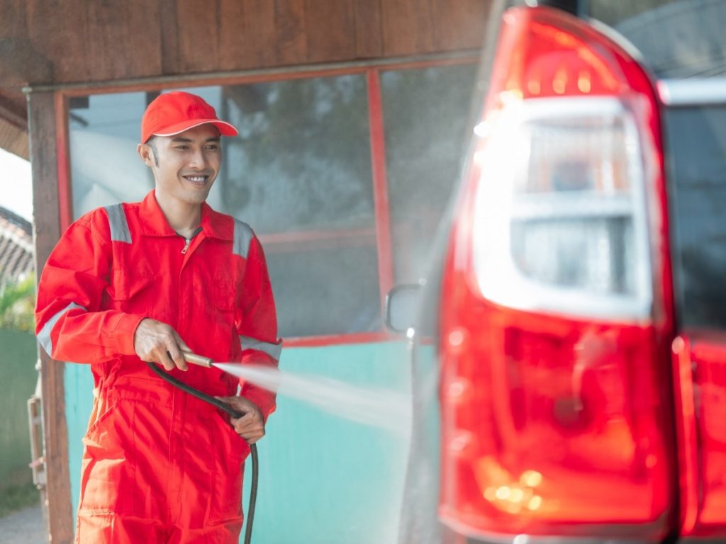 A professional car wash employee in a red jumpsuit smiles while using a pressure washer to clean a dark SUV, promoting Membership Car wash Services in Katy TX.