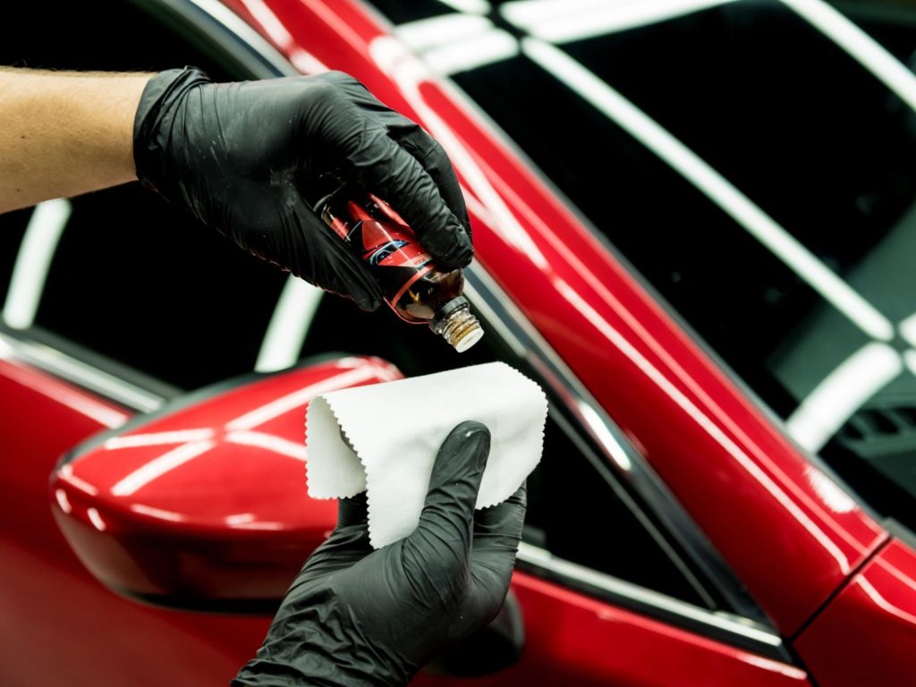 Gloved hand applying premium car detailing product to a microfiber cloth on a red vehicle at Drive-Thru Car Wash Katy TX.