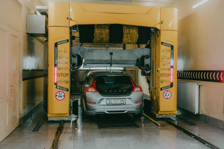 Drive-thru Car Wash Katy TX: silver Volvo in automated tunnel wash with yellow machinery, rotating brushes and nozzles.