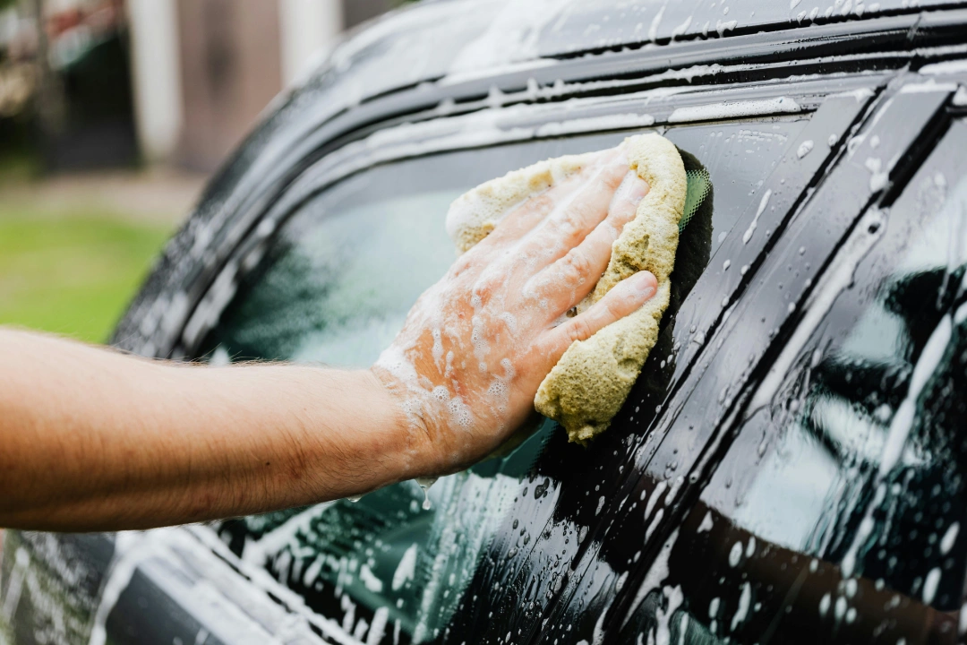 Close-up of a hand washing a black car's side window with a large sponge, offering Affordable Car Wash Katy TX.